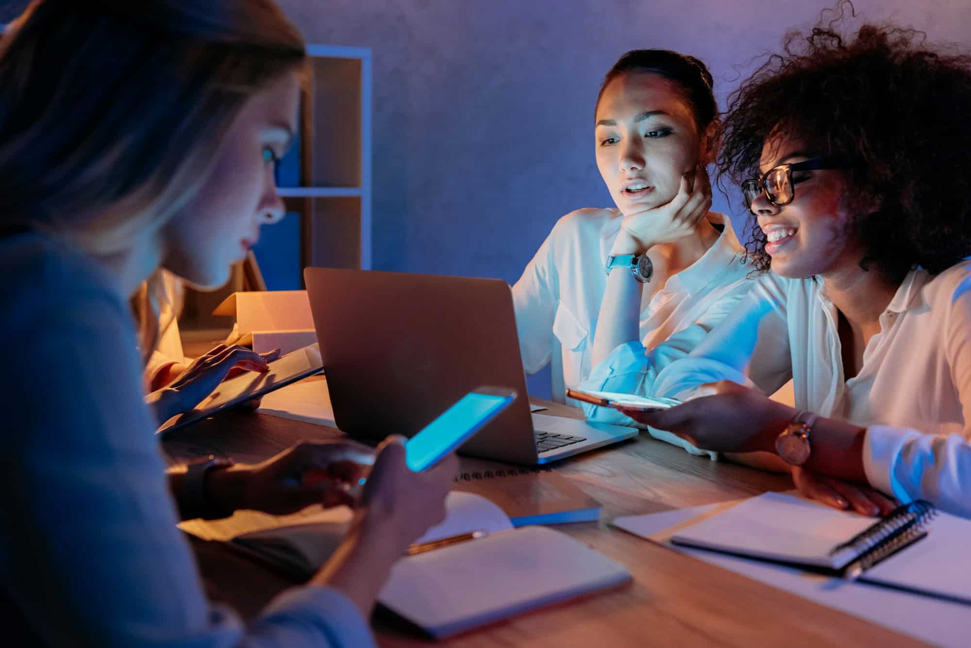 "Three young businesswomen working together on a laptop in a dimly lit office, with glowing screens illuminating their faces."