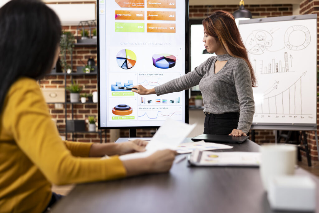 "A woman presenting analysis on a large screen to another person in an office setting."
