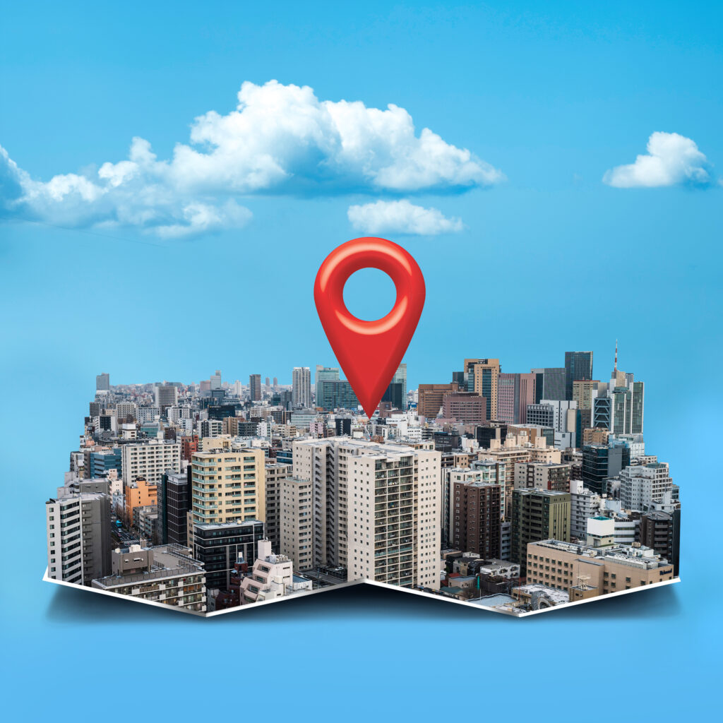 "A red location pin icon stands prominently over a city skyline, set against a clear blue sky with white clouds."
