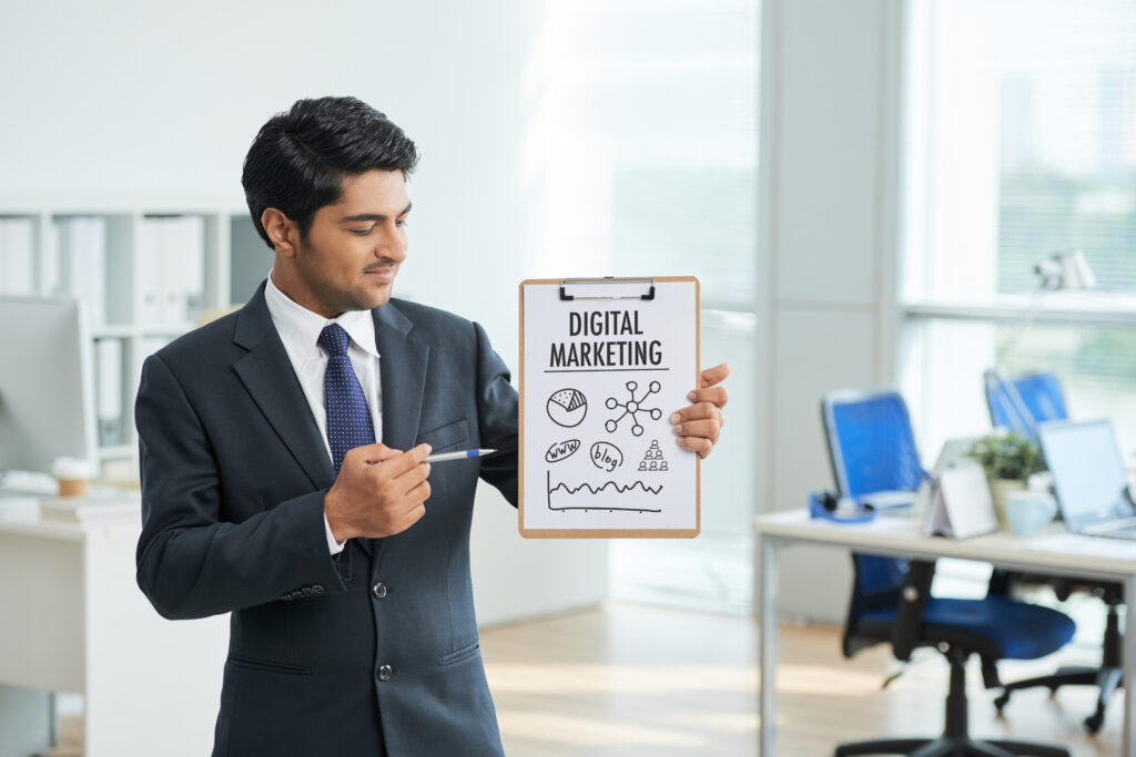 "A man in a suit stands in an office, holding a clipboard with "DIGITAL MARKETING" written on it and pointing to it."